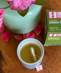 Cup of steeping green tea next to box of Equal Exchange Green Tea and a light green vase with pink flowers 